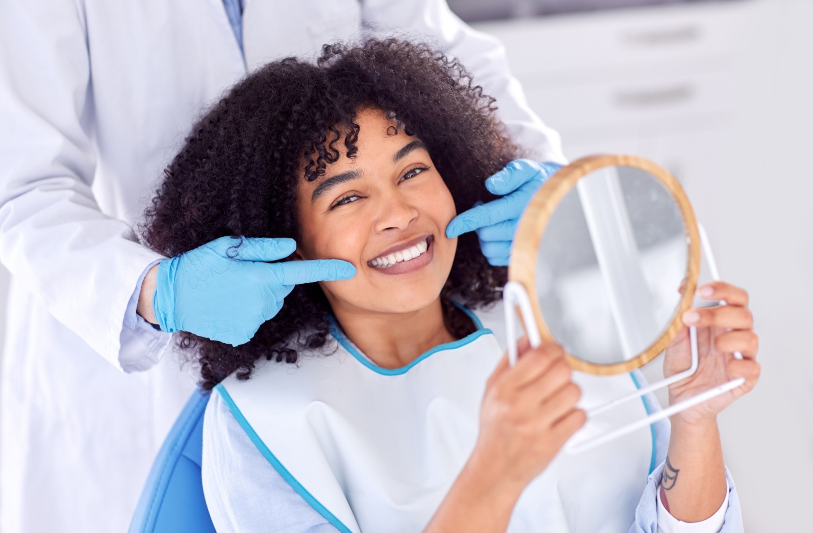 Person smiling while holding mirror during dental checkup as dentist points to their cheeks with gloved hands