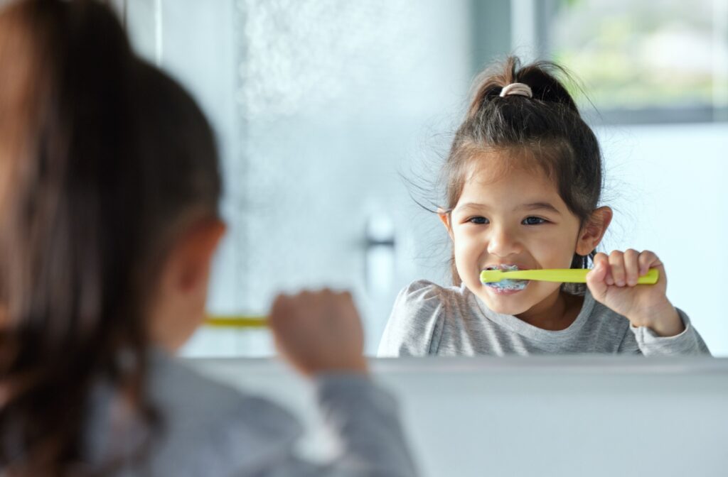 A young child brushing her teeth in the mirror using kid-friendly toothpaste