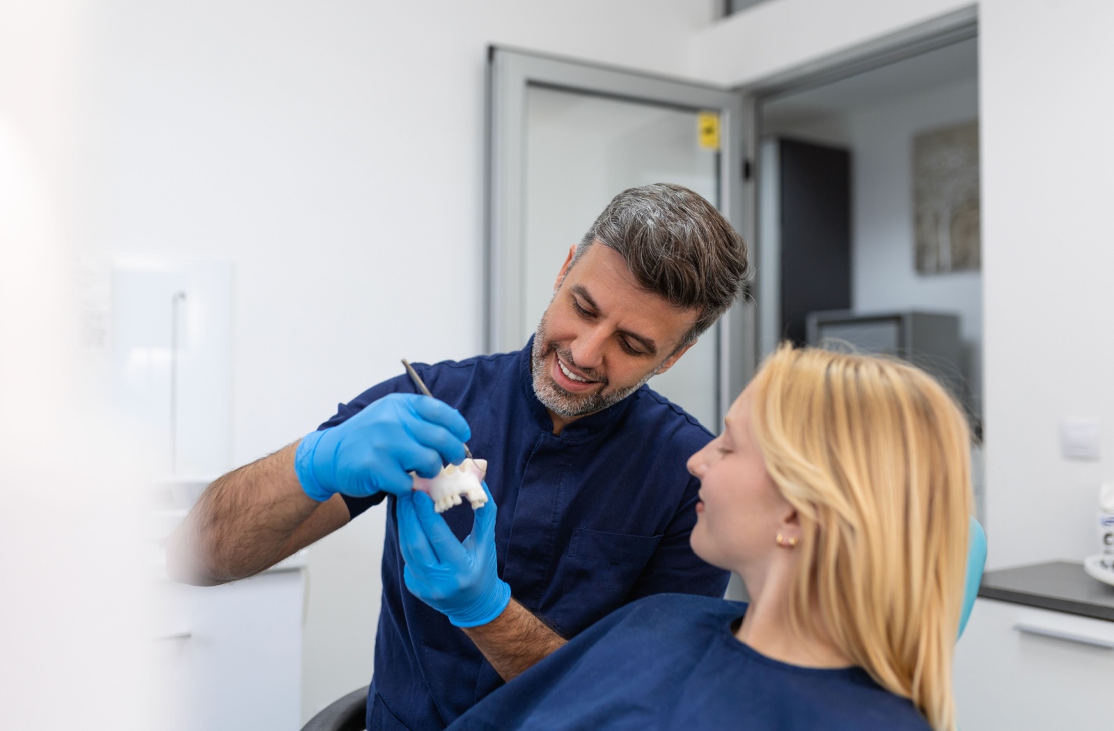 A dentist using a model of a mouth to explain to a patient why they might be experiencing tooth pain under their dental bridge.