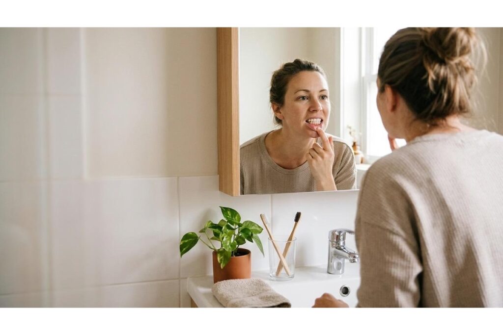 Adult in a bathroom mirror pulling down their lower lip to look at their teeth and gums, beside a sink with two bamboo toothbrushes in a glass